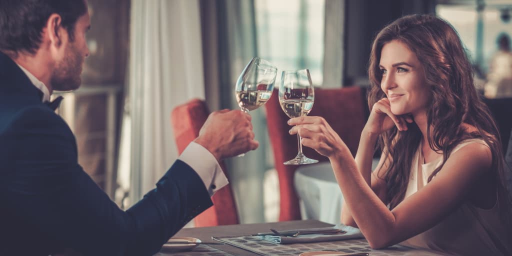 A man and woman smiling and toasting glasses of white wine during a date at an upscale restaurant.