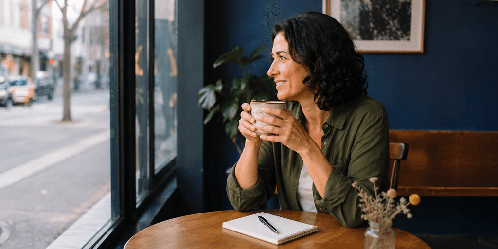 A woman in her 50s sits by a café window holding a mug and smiling thoughtfully, with a notebook on the table beside her, representing the reflective work of rediscovering herself and clarifying what she truly wants in a partner after divorce.