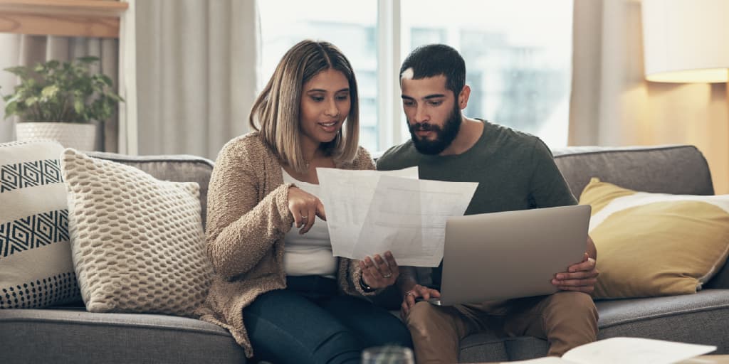 Couple reviewing financial documents together while sitting on a couch with a laptop.