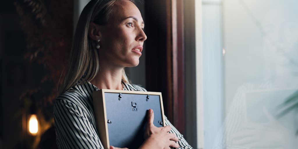 Woman looks out a window while holding a framed photo against her chest, its back facing the viewer.