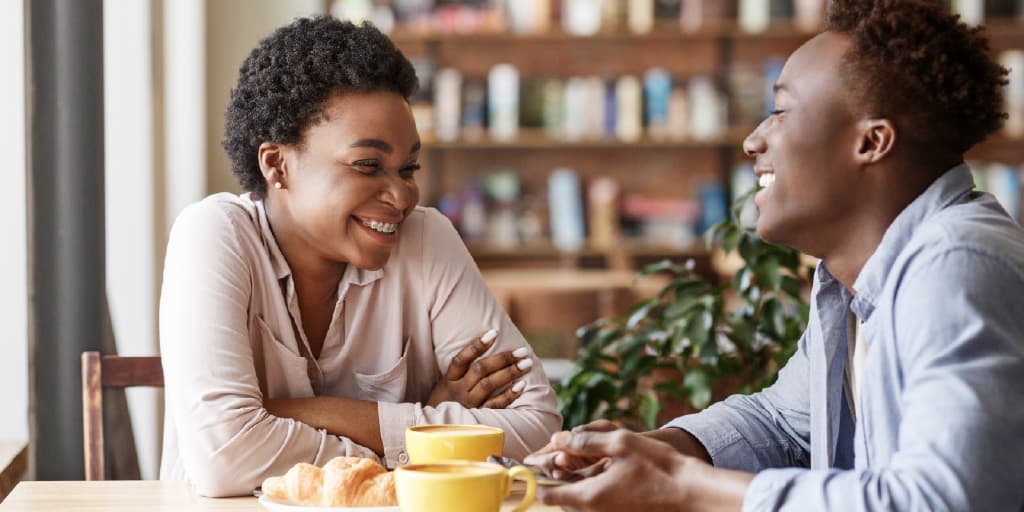 Two people smiling at each other across a cafe table with yellow coffee cups and croissants.