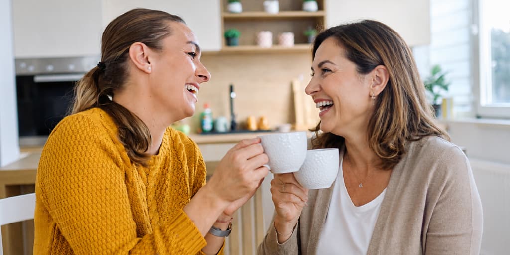 Two women laughing and smiling while clinking white coffee mugs together in a kitchen setting.