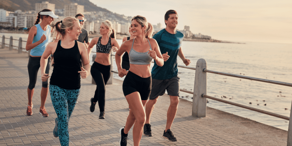 Keep pursuing hobbies and passions. A group of friends in athletic apparel run together on a boardwalk along the coast with a beach city in the background.