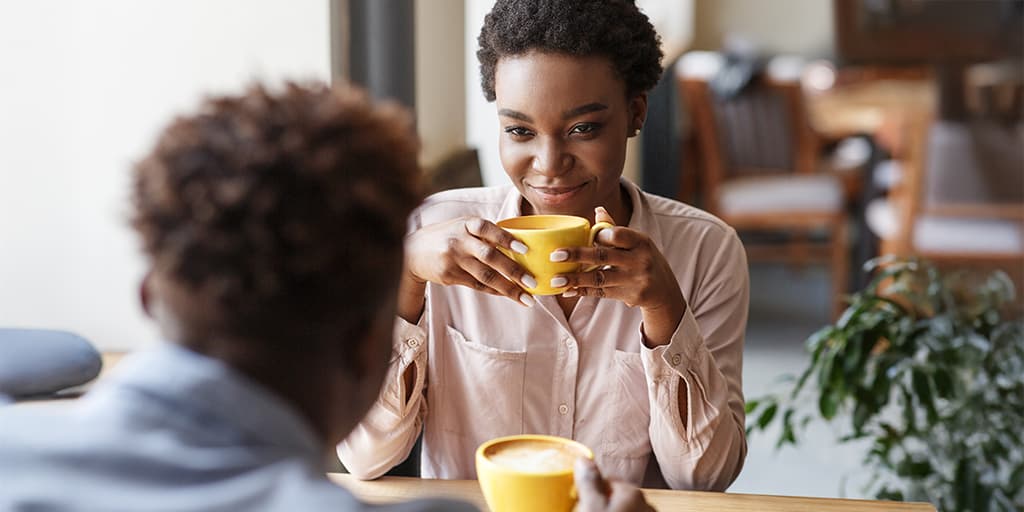 A woman holds a cup of coffee at a cafe smiling at her date who sits across the table.