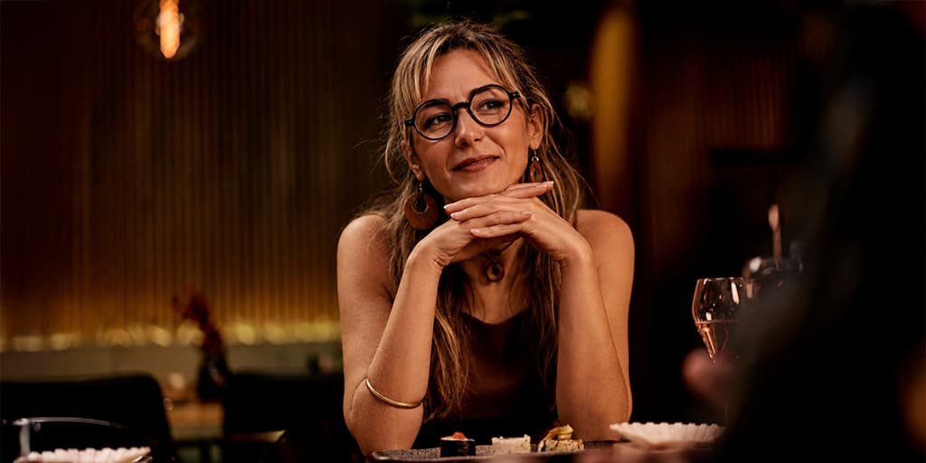 A woman in her 40s with glasses rests her chin on her hands and smiles thoughtfully across a candlelit restaurant table on a first date, representing the relaxed, present version of herself that emerges after doing the eight months of post-divorce work before dating again.