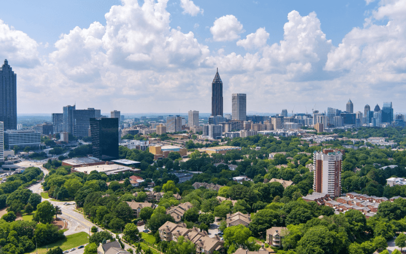 Aerial view of the Atlanta skyline rising above leafy, tree-filled neighborhoods on a sunny day.