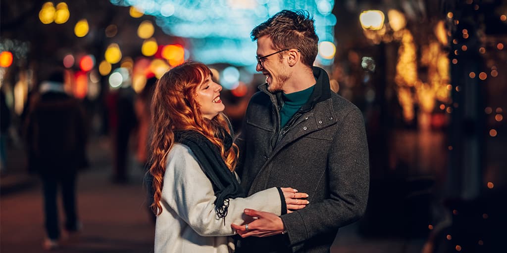 A smiling couple stands close together on a city street at night, lit by warm, glowing string lights and blurred storefronts in Chicago's River North neighborhood.
