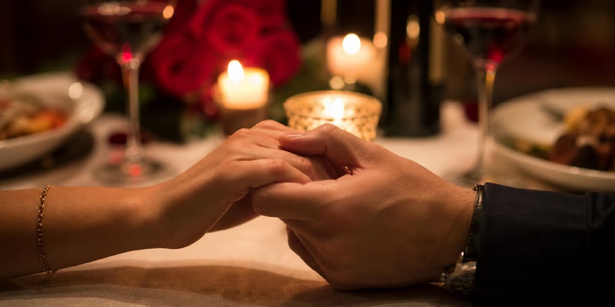 Couple holding hands across a candlelit dinner table set with wine glasses and roses, illustrating a romantic date.