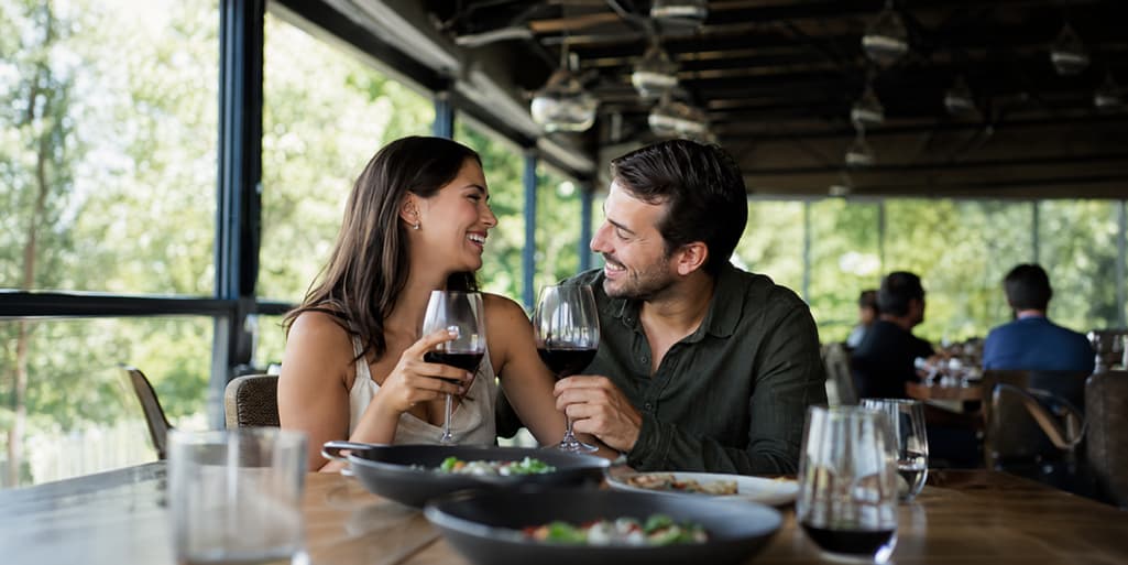 A smiling couple enjoys red wine and shared plates during a dinner date at a bright, upscale restaurant.