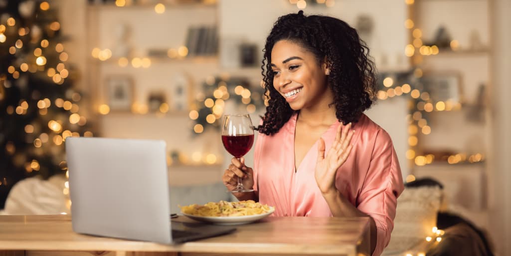 Woman smiling and waving during a video call while holding a glass of wine and sitting at a table with a laptop, suggesting a virtual date or long-distance relationship.