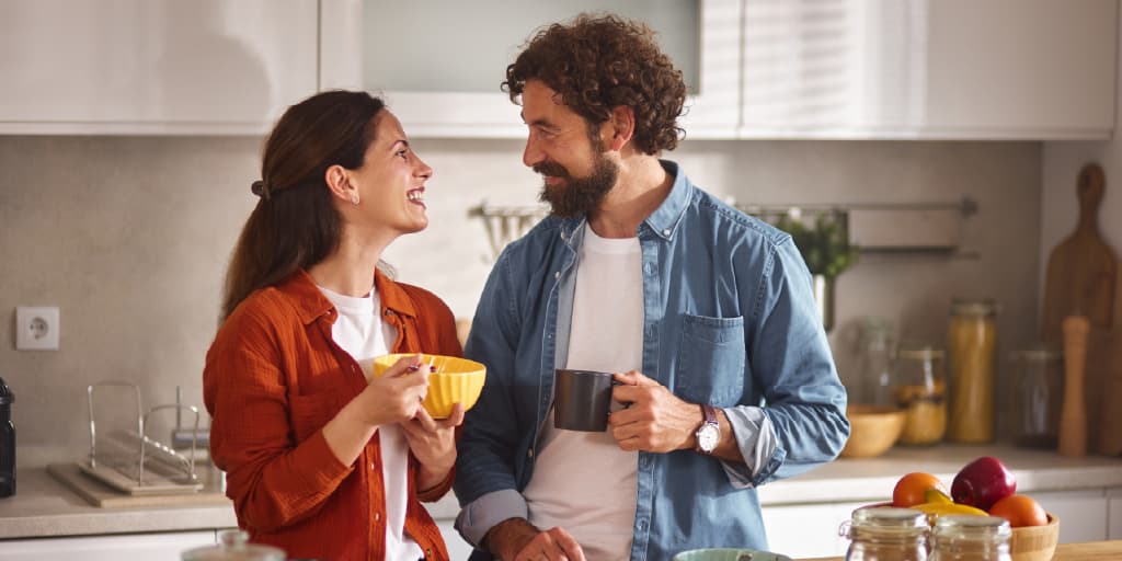 A man wearing a blue jean button shirt and woman wearing a red button shirt smile at each other while sipping coffee in their kitchen.