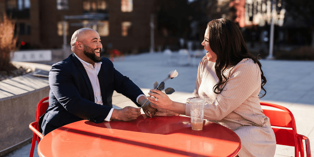 A man in a suit gives a woman a white rose while having a date at a cafe patio table in the city.